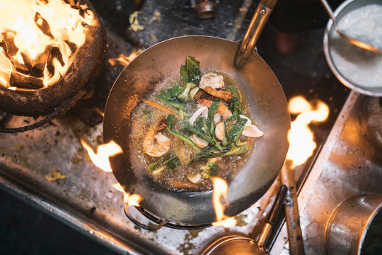 High angle close up of stir fry with prawns and green vegetables in stainless steel wok over flame of charcoal grill.