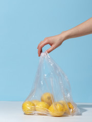 A woman's hand holds a few lemons and a plastic bag on a blue background