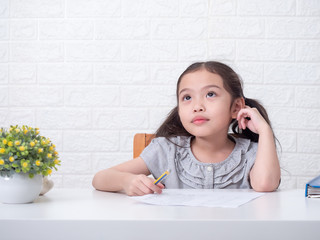 Asian little cute girl 6 years old holding pencil and thinking about math homework over white brick wall background and white table. Learning and education