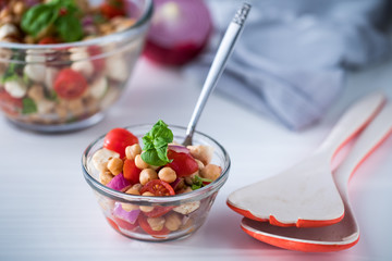 A close up of a small glass bowl filled with chick pea salad with salad tongs.