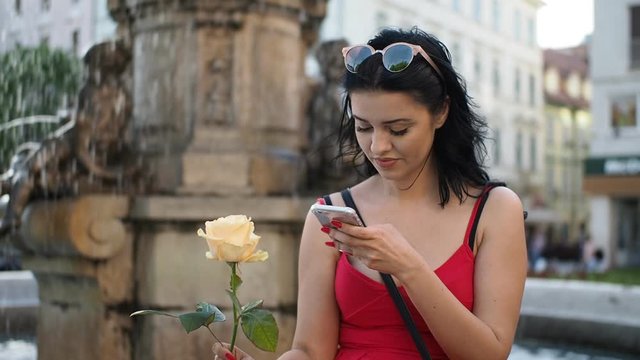 Young enamored woman holding and smelling yellow rose flower and sitting on the fountain in historical part of Bratislava city. She is using smartphone for texting. HD VIDEO.
