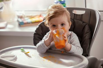 Beautiful baby in the child dining chair eat healthy, vegetable food. Child eat  pumpkin and carrot puree. Lifestyle. Sunny day. Dirty face of happy kid. Portrait of a baby eating with a stained face.