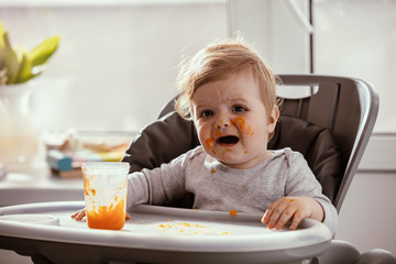 Beautiful baby in the child dining chair eat healthy, vegetable food. Child eat  pumpkin and carrot puree. Lifestyle. Sunny day. Dirty face of happy kid. Portrait of a baby eating with a stained face.