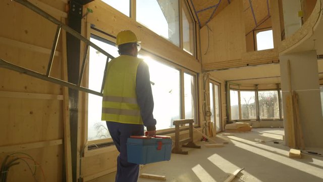 SLOW MOTION, LENS FLARE, CLOSE UP: Golden sunbeams shine on worker coming to work on a contemporary prefabricated wooden house. Unrecognizable builder arrives to work carrying his toolbox and ladder.