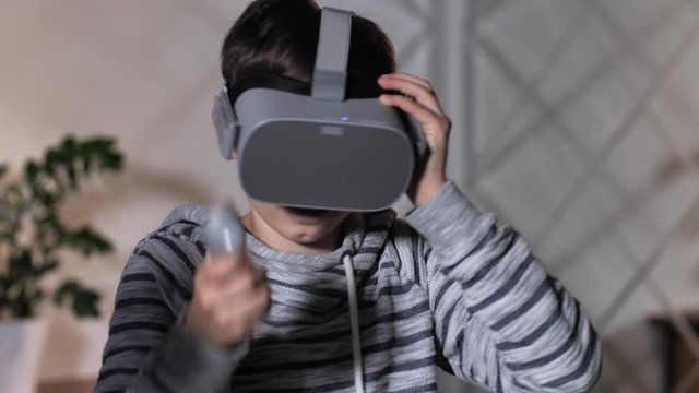 Close-up of modern teenager in striped hoodie wearing VR goggles and using joystick while going to play video game at home. Teenage boy preparing to dive into game world with augmented-reality headset