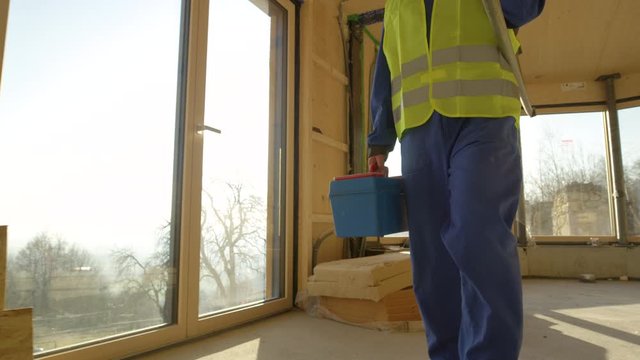 SLOW MOTION, CLOSE UP, LENS FLARE: Unrecognizable builder in blue coveralls leaves the beautiful wooden prefabricated house on a sunny evening. Contractor arrives to work in a cross-laminated house.