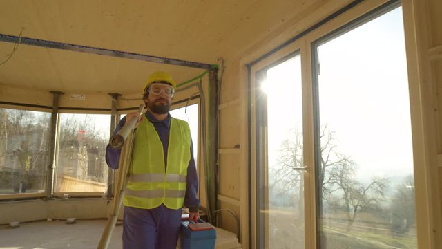 SLOW MOTION, LENS FLARE, CLOSE UP: Caucasian man in blue coveralls and yellow hard hat leaves the construction site on a sunny afternoon. Builder arrives to work in a beautiful contemporary log house.