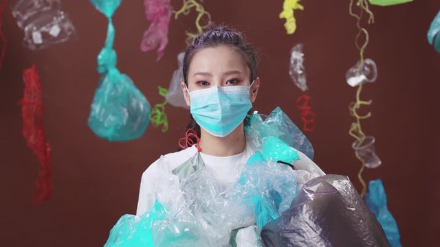 Female eco activist wearing a protective mask and dangerous plastic packaging dress look at camera on background. Save the planet. Overconsumption concept.
