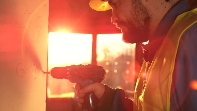 SLOW MOTION CLOSE UP LENS FLARE, DOF: Caucasian builder unscrews a bolt stuck in a wall panel. Contractor working at sunrise uses his electric drill to unfasten a screw holding together drywall panels