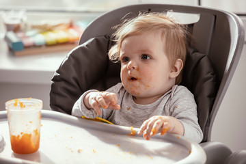  Baby in the child dining chair eat healthy, vegetable food. Child eat  pumpkin and carrot puree with banana. Sunny day. Dirty face of happy kid. Portrait of a baby eating with a stained face.