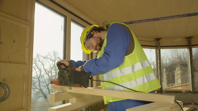 SLOW MOTION, LENS FLARE, CLOSE UP, DOF: Young Caucasian man working on a prefabricated house buffs a plank with a sander. Builder wearing a helmet and yellow vest sands a wooden beam on a sunny day.