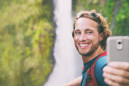 Selfie travel tourist man taking photo with phone on adventure hike vlogging for online social media influencer. Hawaii waterfall sightseeing. Smiling backpacker guy using mobile on summer vacation.
