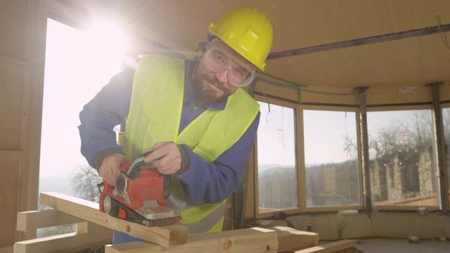 SLOW MOTION, PORTRAIT, LENS FLARE: Caucasian construction worker smiles while sanding a wooden beam on a sunny day. Happy contractor building a contemporary hardwood house smoothens a wooden board.