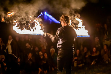 A man is seen holding burning swords during a fire dance routine by night, with blurry people...