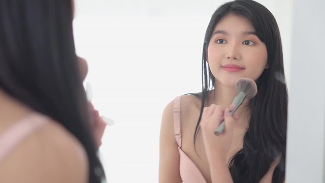 Beauty young asian woman happy and smile with face looking mirror applying makeup with brusher of cheek in the room, beautiful of girl holding brush make-up on facial, skin care and cosmetic concept.
