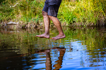 A healthy man wearing shorts with soaked legs is seen balancing on a slackline over a pond in nature, barefooted and close to surface with reflection