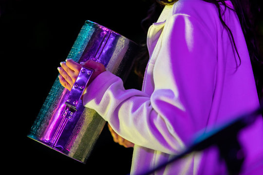 A close up and side view of a musician using a metal tin can made into a traditional percussion instrument, with violet stage lighting at a festival