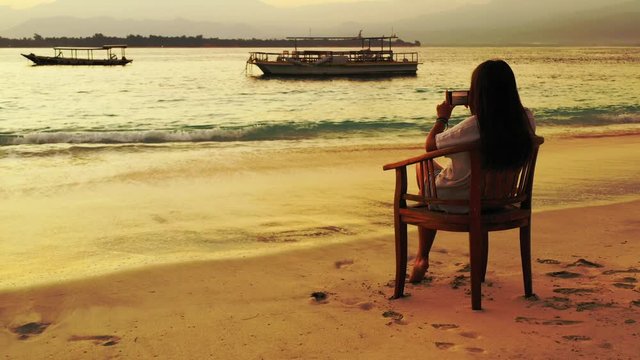 Young woman sitting on exotic beach taking pictures of beautiful calm lagoon with anchored boats at sunset golden hour in Philippines