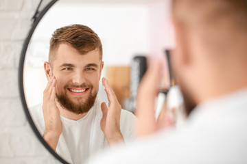 Handsome young man applying cream on his face near mirror in bathroom - Powered by Adobe