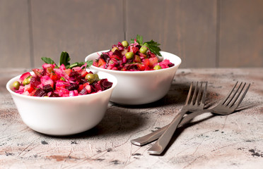 Still life. On a brown table, against a background of a gray wooden wall, are white bowls with a bright red vegetable salad decorated with a green leaf of parsley. Nearby are two metal forks. Cooking,