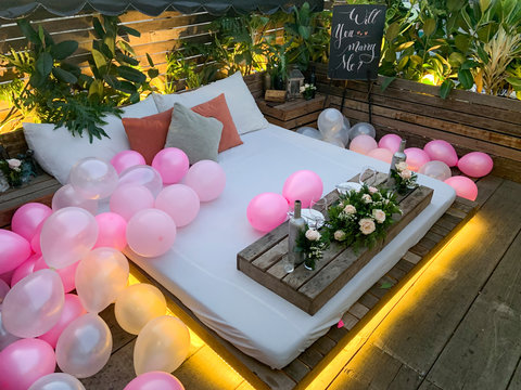 A Sign Reads Will You Marry Me. A Romantic Surprise Outdoor Garden Setting With Bed, Colored Balloons And Table For Two With A Pink Theme