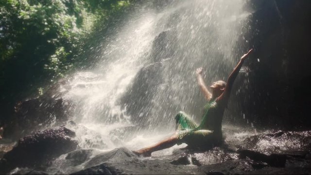 young attractive Asian Indonesian woman in sarong overwhelmed by the beauty of waterfall in Bali tropical jungle letting water falling on her feeling free and blissful