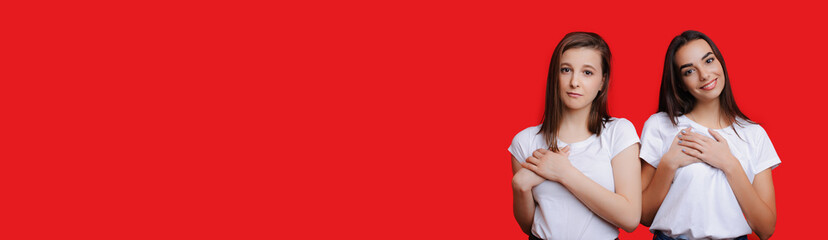 Two charming caucasian girls with black hair and beautiful smile are touching their hearts while advertising something on red freespaced background