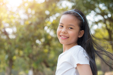 photo happy little asian girl child standing with big smile..fresh healthy green bio background with abstract blurred foliage and bright summer sunlight. concept of happy family or successful adoption