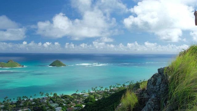 Young woman stands on the hill and takes picture of the windward coast of Oahu Island with Mokulua Islands on the background