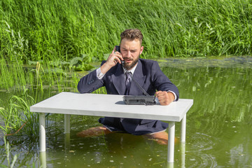 A man in a jacket and tie sits without pants at a white table in a swamp and holds a telephone...