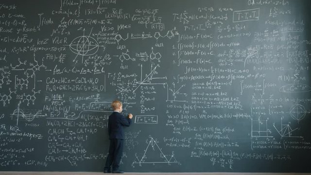 Prodigy child in suit is writing formulas on huge blackboard in school classroom concentrated on educational activity. Education and childhood concept.
