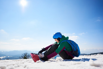 Female snowboarder wearing colorful clothing and helmet with backpack sitting in snow, adjusting snowboard on copy space background of blue sky and winter mountain landscape - snowboarding concept.