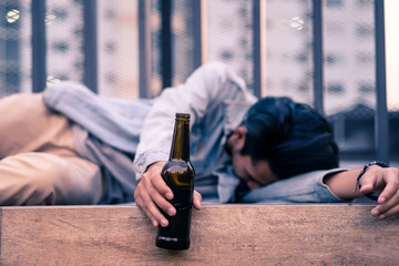 Asian drunk man holding beer bottle lying on floor. Young male adult crying having problem and feeling stress then drinking beer to forget those trouble and sleeping on ground. Drunk unhealthy concept