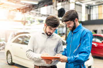 Vehicle maintenance handsome man servicer explain car condition to male customer in garage. Automotive mechanic pointing needed for the on automotive checklist document. Car repair service concept. - Powered by Adobe