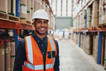 Happy male supervisor in warehouse standing in uniform with white hardhat smiling looking at camera...