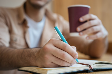 Close up of male hands taking notes on notebook, holding coffee cup. Business man working project in office, focus on hand 