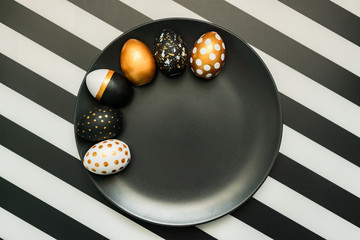 Black and white striped background with white, black and golden decorated eggs on black plate. Trendy easter flat lay.