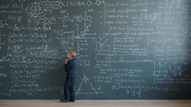 Thoughtful kid in suit is walking near board with formulas in school class thinking then going away. Intelligent children, modern education and lifestyle concept.