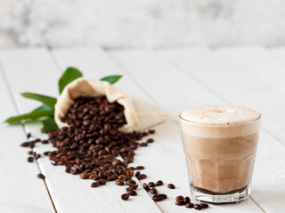 Cappuccino or latte with milk froth in a glass. Сoffee beans in a bag of coffee and coffee leaves on a white wooden background. Soft focus, close up.