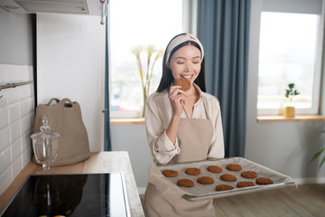 Young girl with baking sheet holding one cookie near mouth.