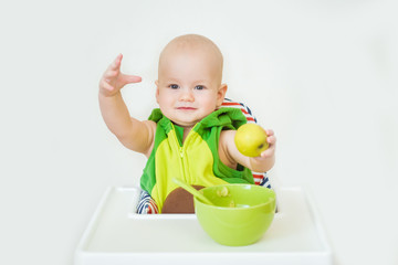 Little happy baby with spoon sits at highchair and eats porridge on plate