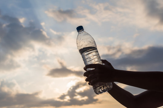 Girl Holding In The Hand A Plastic Bottle With Crystal Clear Mineral Water Opposite The Rays Of The Sunset Sky. Concept