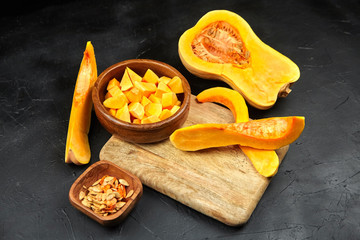 Butternut squash pieces, wooden bowl with pumpkin seeds, cutting board on black background, closeup. Cooking winter squash
