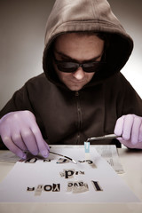 Criminal in hooded shirt preparing ransom letter from newspaper