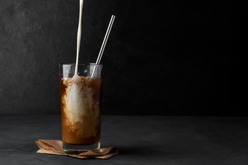 Iced coffee with pouring cream in a glass cup with a metal straw on a dark background