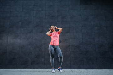 Fototapeta na wymiar Full length of attractive smiling Caucasian brunette in sportswear and with ponytail listening music over headphones. In background is dark wall.