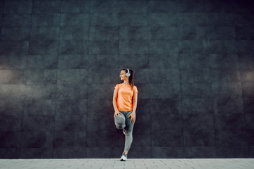 Fototapeta premium Full length of attractive smiling Caucasian brunette in sportswear and with ponytail listening music over headphones while leaning on the wall. In background is dark wall.