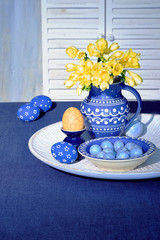 Easter decorations in classic blue and yellow colors. Orange egg with polka dots and yellow freesia flower, traditional ceramic vase, jug and plate on blue linen tablecloth. Interior Spring design.