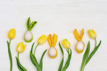 Easter composition with handmade colorful easter eggs with ears from rabbit and fresh yellow flowers tulip on white wooden background with copy space. View from above, flat lay.