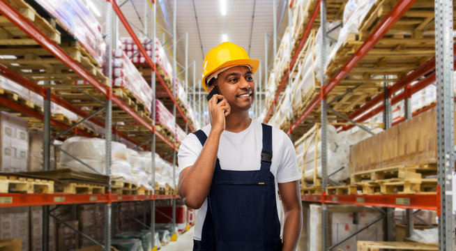 profession, logistic business and technology concept - happy smiling indian worker or loader in helmet calling on smartphone over warehouse background